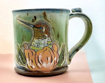 May include: A ceramic mug with a light green glaze featuring a hand-painted hummingbird with orange and white plumage perched inside a large, stylized orange flower. The mug has a sturdy handle and a dark brown base with blue rim accents.