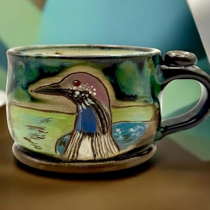 May include: A ceramic mug featuring a hand-painted illustration of a loon on a lake. The mug has a glossy finish with shades of green, blue, and brown. It includes a dark handle and a small, rounded thumb rest on top of the handle for grip.