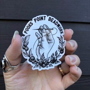 May include: A white sticker featuring the text "POOKS POINT BEADWORK" around a black and white illustration of a hand threading a needle. The design is framed by a wreath of leaves and berries.