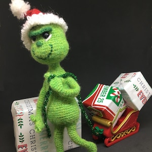 May include: A green crocheted Grinch doll wearing a red and white Santa hat, pulling a red sleigh with wrapped presents. The presents are wrapped in white paper with green and red designs and the text "Christmas Eve" and "December 25".