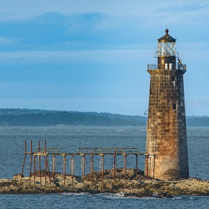 May include: A weathered stone lighthouse stands on a rocky islet in the ocean under a blue sky. The lighthouse has a square base and a lantern room at the top. A wooden pier extends from the islet. The scene evokes a sense of maritime history and coastal beauty.