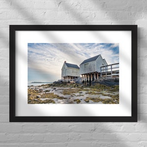 May include: Two white wooden houses on stilts stand on a rocky shore with seaweed and tide pools. The sky is cloudy and blue.