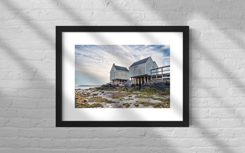 May include: Two white wooden houses on stilts stand on a rocky shore with seaweed and tide pools. The sky is cloudy and blue.