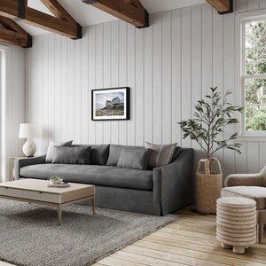 May include: A living room with a gray sofa, a coffee table, a chair, and a framed photograph on the wall. The room has a wooden floor and white walls.