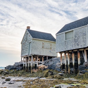 May include: Two weathered, gray wooden houses on stilts stand on a rocky shore. The houses have a rustic charm and are surrounded by water and rocks.