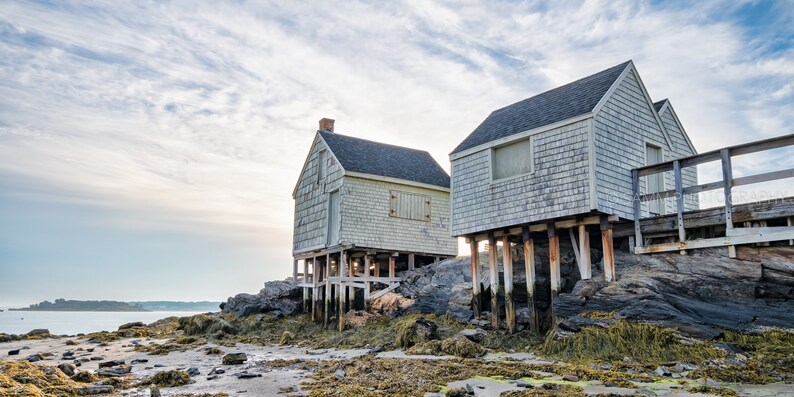 May include: Two weathered, gray wooden houses on stilts stand on a rocky shore. The houses have a rustic charm and are surrounded by water and rocks.
