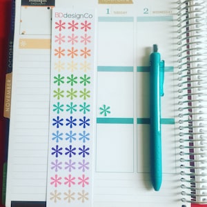 May include: A planner open to November with a teal pen and a sticker sheet from BDdesignCo. The sticker sheet has colorful asterisks. The planner has a spiral binding and is on a red surface.