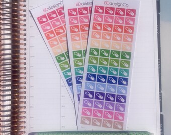May include: Three colorful sticker sheets with pill icons, labeled "BDdesignCo," rest on an open planner. A green pen with a leaf pattern lies below. The planner has a spiral binding and a weekly layout.