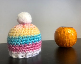 May include: A colorful crocheted hat with a white pom pom. The hat is striped with pink, blue, yellow, and orange yarn. The hat is sitting on a dark surface next to a round, orange object.