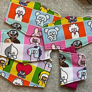 May include: A set of six colorful fabric face masks featuring various cartoon characters. The masks are made of cotton and have a variety of designs, including a heart, a camera, and a bunny. The masks are all different colors, including pink, blue, yellow, and green.