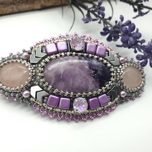 Peut inclure: Broche ovale ornée d'une pierre centrale violette, entourée de perles roses, argentées et violettes. Le design comprend des accents métalliques argentés et des cristaux transparents. La broche est finement détaillée, mettant en valeur textures et couleurs.