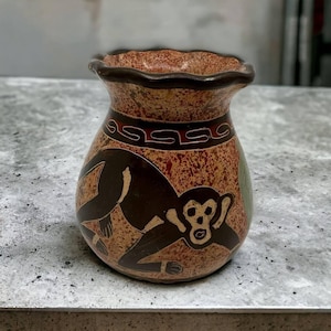 May include: A small, handcrafted vase with a brown and tan color scheme. The vase features a dark brown monkey illustration and decorative patterns. The rim is dark brown, and the vase has a unique, textured appearance.