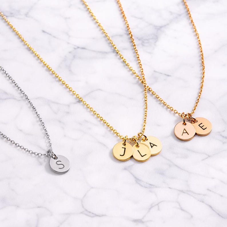 Three dainty initial disc necklaces in silver, gold, and rose gold finishes displayed on a white marble background