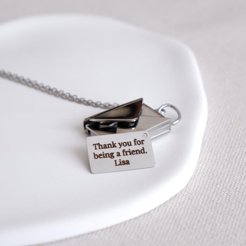 May include: A silver-toned necklace featuring an envelope pendant with a small tag. The tag is engraved with the message "Thank you for being a friend. Lisa". The necklace is displayed on a white surface.