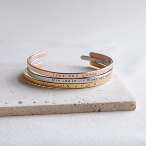 May include: Three open cuff bracelets in gold, rose gold, and silver. The bracelets are engraved with the phrases "Jack and Amber", "I love you to the moon and back", and coordinates 51° 30' 26" N 0° 7' 40" W.