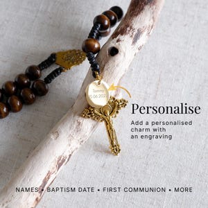 May include: A rosary with dark wooden beads and a gold-colored crucifix. A round, gold-colored charm is engraved with the name "Tommy" and the date "15.06.2022". The image includes the text "Personalise" and "Add a personalised charm with an engraving".