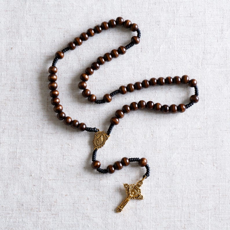 May include: A rosary with dark brown wooden beads and black cord. The rosary features a gold-colored crucifix and a gold-colored centerpiece with a religious image. The beads are evenly spaced along the cord.