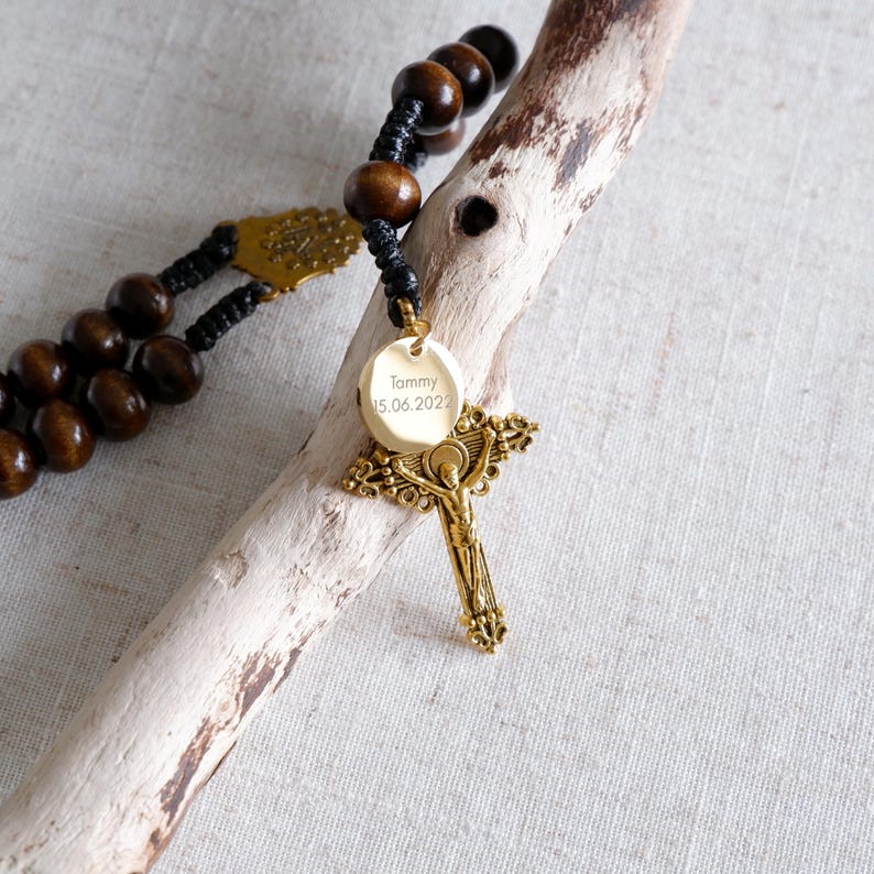 May include: A rosary with dark brown wooden beads and a gold-colored crucifix pendant. The pendant has the name "Tommy" and the date "15.06.2022" engraved on it. The rosary is resting on a piece of light-colored driftwood.