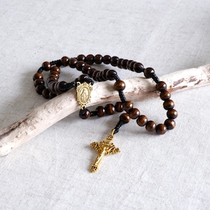May include: A rosary with dark brown wooden beads and a gold-colored crucifix. The beads are strung on black cord, with a gold-colored centerpiece featuring a religious image. The rosary is displayed on a light-colored surface.