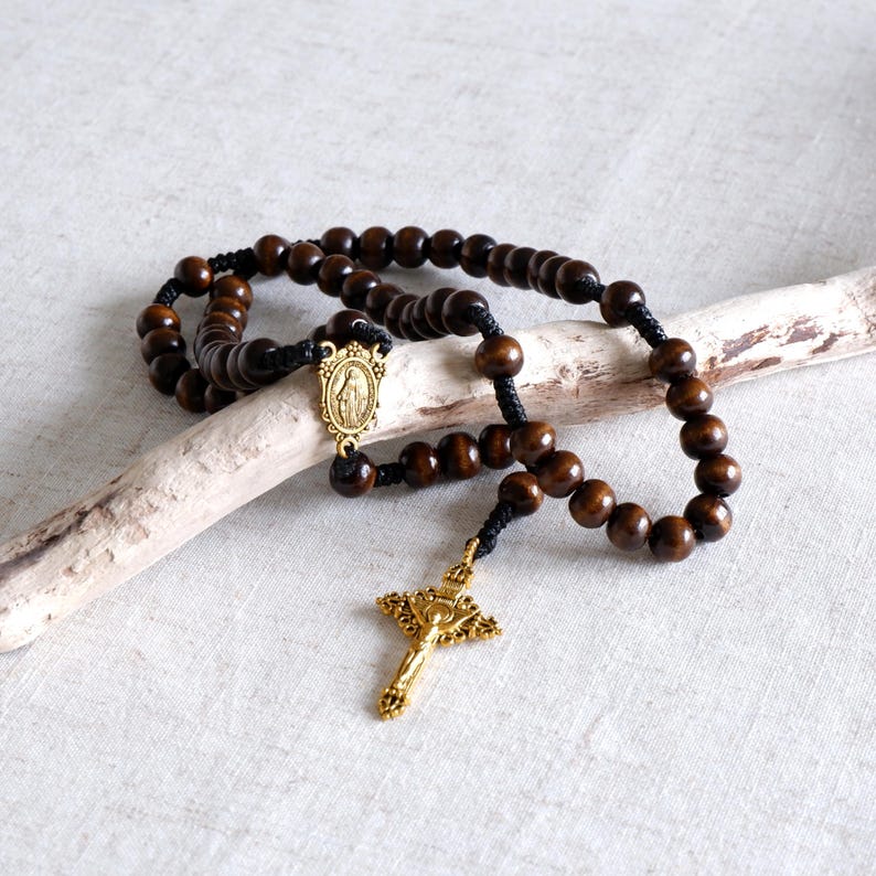 May include: A rosary with dark brown wooden beads and a gold-colored crucifix. The beads are strung on black cord, with a gold-colored centerpiece featuring a religious image. The rosary is displayed on a light-colored surface.