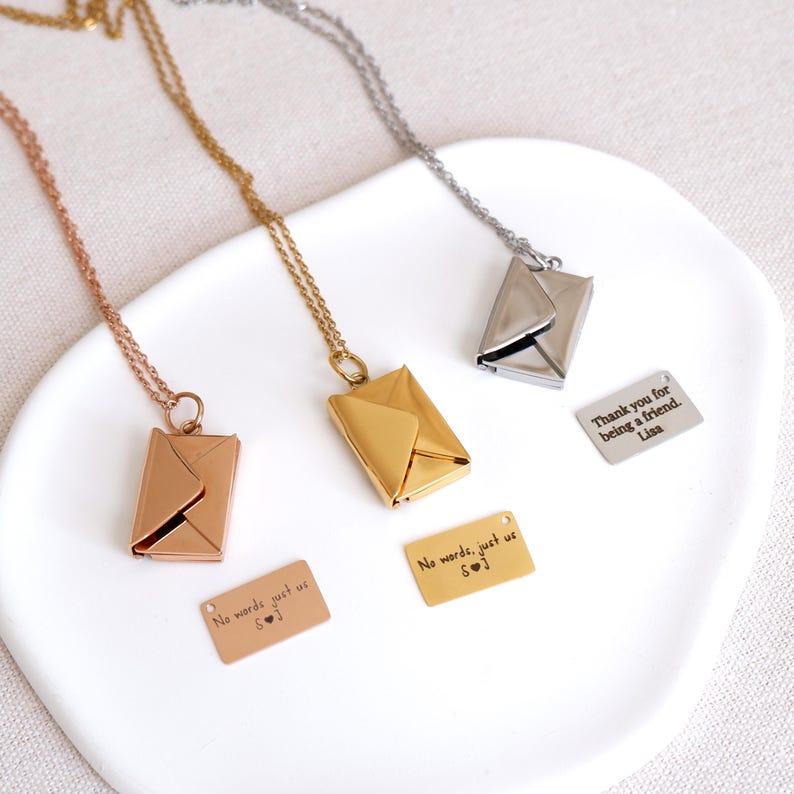 May include: Three envelope pendant necklaces in rose gold, gold, and silver. Each necklace has a small rectangular tag with text. One tag reads "No words, just us S&hearts;J", another reads "Thank you for being a friend. Lisa".