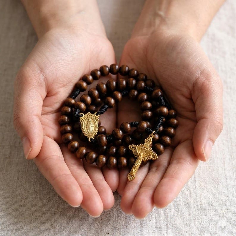 May include: A wooden rosary with dark brown beads and gold-colored metal accents, including a crucifix and a medallion. The rosary is held in open hands, showcasing its details.