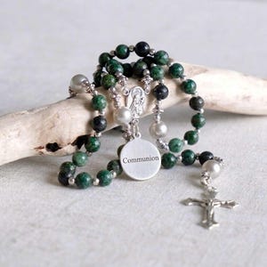 Peut inclure: Un chapelet avec des perles vertes et noires, des accents argentés et un pendentif en forme de croix. Le chapelet comporte un charm de communion. Les perles sont faites d'une pierre tachetée. Le chapelet est exposé sur une surface claire.