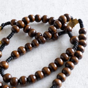 May include: A close-up of a rosary with dark brown wooden beads. The beads are strung on black cord, with a gold-colored metal cross at the end. The beads are smooth and round, and the cord is tightly woven.