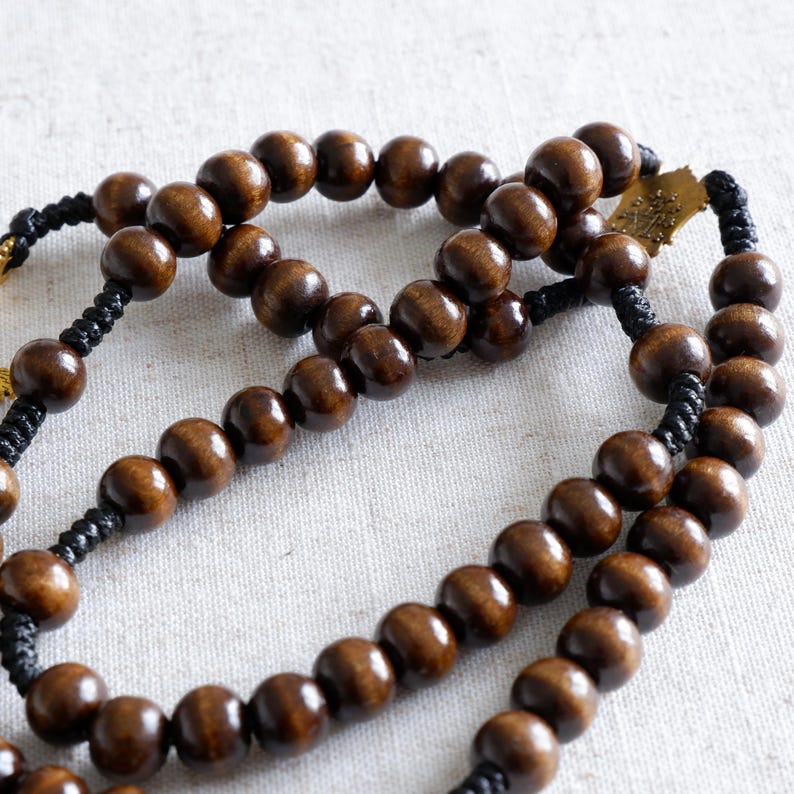 May include: A close-up of a rosary with dark brown wooden beads. The beads are strung on black cord, with a gold-colored metal cross at the end. The beads are smooth and round, and the cord is tightly woven.