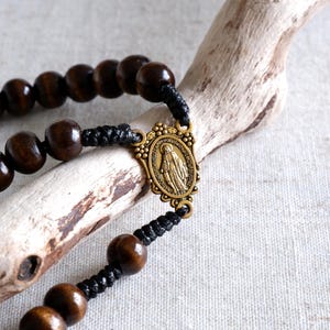 May include: A rosary with dark brown wooden beads and a bronze-colored centerpiece featuring a religious figure. The beads are strung on black cord. The rosary is displayed on a piece of driftwood.