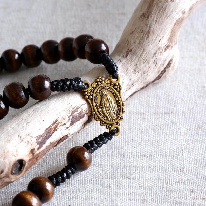May include: A rosary with dark brown wooden beads and a bronze-colored centerpiece featuring a religious figure. The beads are strung on black cord. The rosary is displayed on a piece of driftwood.