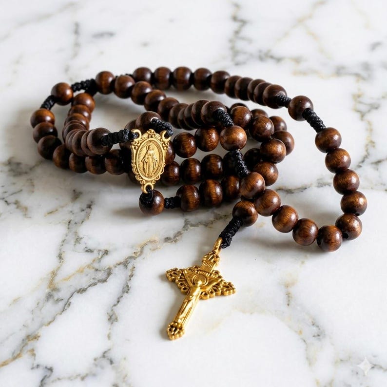 May include: Wooden rosary with dark brown beads and black string. The rosary features a gold-colored crucifix and a gold-colored centerpiece with a religious figure. The beads are round and evenly spaced.