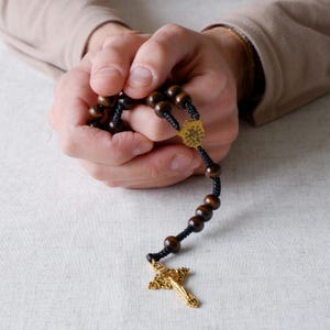 May include: A rosary with dark brown wooden beads and a gold-colored crucifix. The beads are strung on black cord, with a gold-colored centerpiece. The crucifix has intricate detailing.