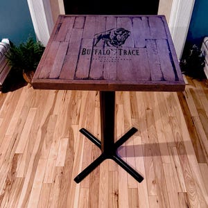 May include: Square wooden table with a dark brown top featuring the "Buffalo Trace" logo and a bison illustration. The table has a black metal base and is set on a hardwood floor.