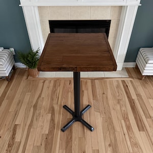 May include: A square, dark brown wooden table with a black metal base. The tabletop is made of planks and is supported by a single, black metal pole. The base has a four-point star design. The table is approximately 30 inches tall.