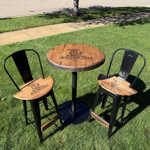 May include: A small round table and two matching chairs with black metal frames and wooden seats and tabletop. The tabletop and seats have a wood grain finish and the text "Mike's Plumbing" is printed on them. The table has a black metal base.
