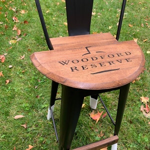 May include: Black metal bar stool with a wooden seat featuring the Woodford Reserve logo. The stool has white protective covers on the legs.
