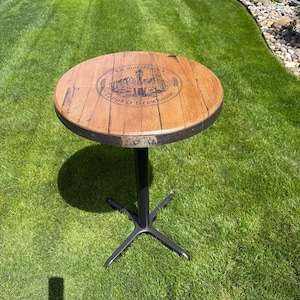 May include: A round, wooden bar table with a dark metal base. The tabletop is made of reclaimed wood with a circular logo that reads "Ed and Mary's Tapped Brewhouse". The table is set on a green lawn.
