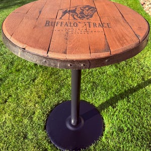May include: A round wooden table made from reclaimed bourbon barrel staves. The tabletop features the "Buffalo Trace" logo and text. The table has a dark metal rim, a single black pedestal, and a round base. The table is set on a green lawn.