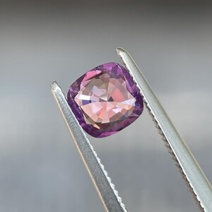 May include: A cushion-cut gemstone held by tweezers. The gem has a gradient of colors, transitioning from a deep purple at the edges to a soft pink in the center. The facets reflect light, creating a sparkling effect. The tweezers are silver.