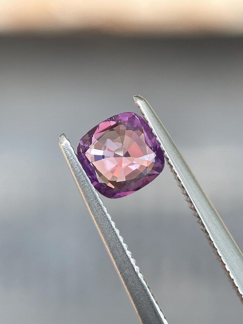 May include: A cushion-cut gemstone held by tweezers. The gem has a gradient of colors, transitioning from a deep purple at the edges to a soft pink in the center. The facets reflect light, creating a sparkling effect. The tweezers are silver.