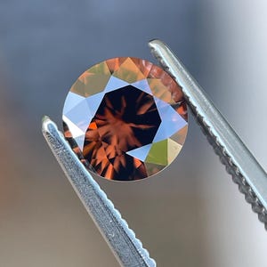 May include: A round, faceted gemstone with a deep reddish-brown color and high clarity. The stone features a brilliant cut with multiple triangular and kite-shaped facets that reflect light, creating a complex pattern of dark and light tones.
