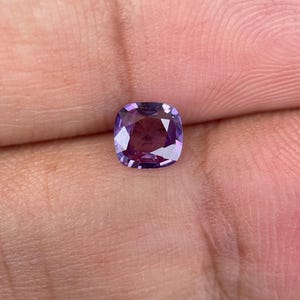 May include: A cushion-cut gemstone with a deep purple hue, held between fingers. The gem's facets reflect light, creating a sparkling effect. The stone's color transitions from a lighter lavender to a darker, richer purple.