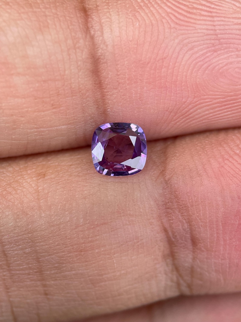 May include: A cushion-cut gemstone with a deep purple hue, held between fingers. The gem's facets reflect light, creating a sparkling effect. The stone's color transitions from a lighter lavender to a darker, richer purple.