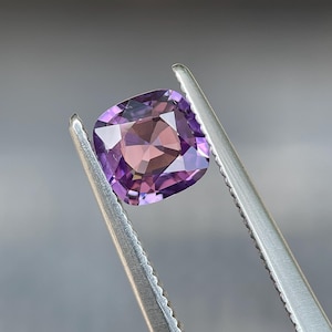 May include: A cushion-cut gemstone held by tweezers. The gem features a gradient of purple and pink hues, with a square shape and faceted surface. The tweezers are silver and the background is a blurred gray.