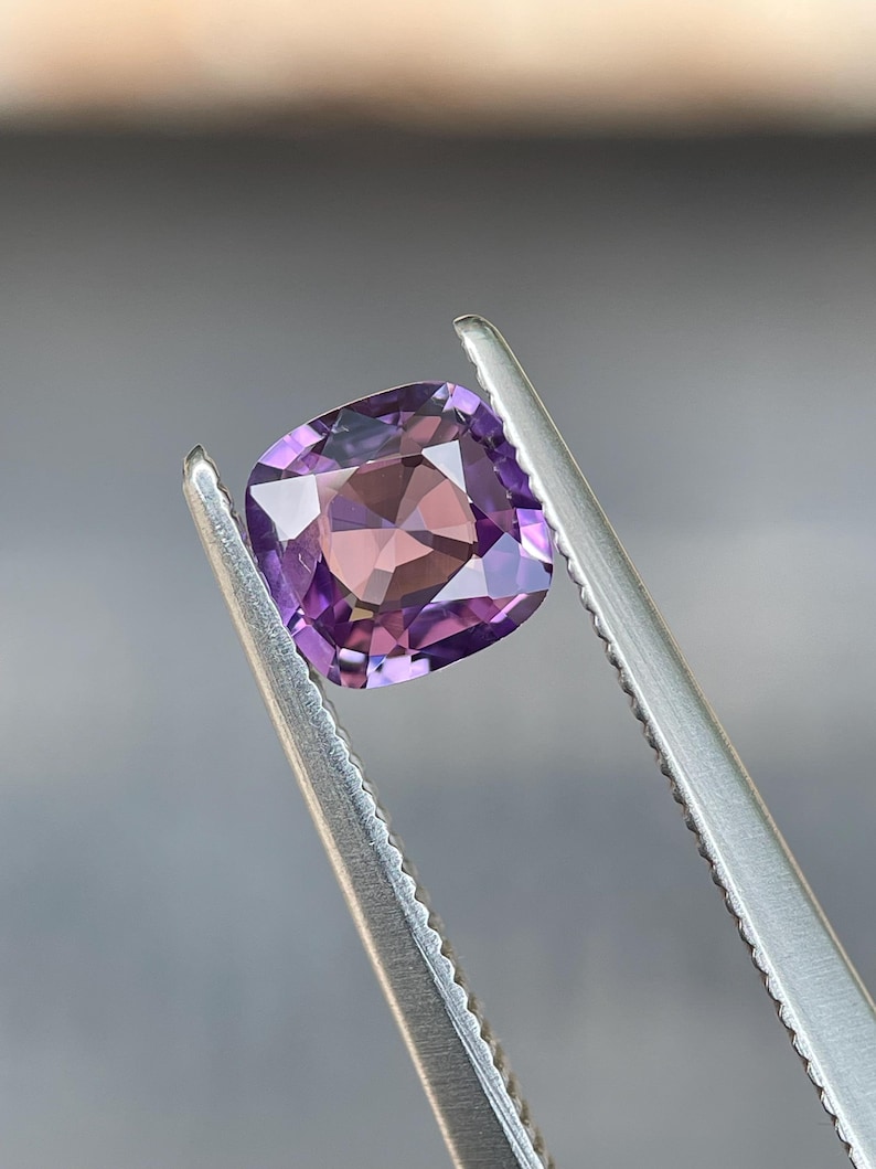 May include: A cushion-cut gemstone held by tweezers. The gem features a gradient of purple and pink hues, with a square shape and faceted surface. The tweezers are silver and the background is a blurred gray.