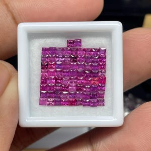 May include: A collection of small, square-cut, deep pink gemstones arranged in a white tray. The gemstones are tightly packed together, creating a mosaic-like appearance. The tray is held by a person.
