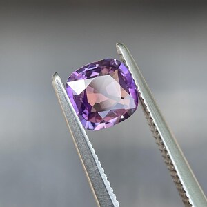 May include: A cushion-cut gemstone, held by tweezers, displays a gradient of purple and pink hues. The gem's facets reflect light, creating a sparkling effect. The tweezers are silver-colored, and the background is a blurred neutral tone.