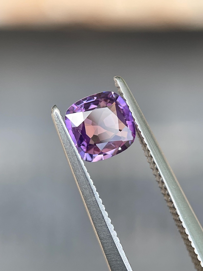 May include: A cushion-cut gemstone, held by tweezers, displays a gradient of purple and pink hues. The gem's facets reflect light, creating a sparkling effect. The tweezers are silver-colored, and the background is a blurred neutral tone.