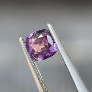 May include: A cushion-cut gemstone with a blend of purple and pink hues is held by a pair of silver tweezers. The gem's facets reflect light, creating a sparkling effect. The background is a soft, blurred gray.